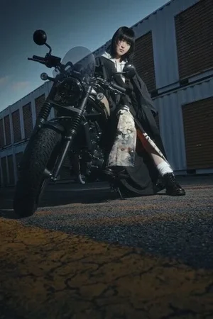 和柄の布を切り替えた黒のロングコートを着て、バイクにまたがるモデル