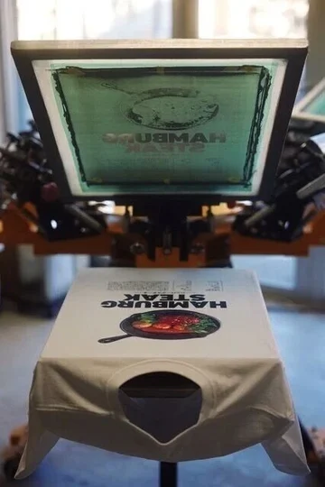スクリーンプリント機で、白いTシャツに「HAMBURG STEAK」のデザインを刷っているところ