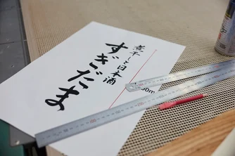 『すぎだま』のロゴ原稿の横に定規と赤鉛筆を置き、看板製作のサイズを検討している様子