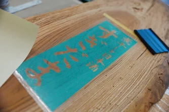 看板となる一枚板に『すぎだま』のロゴを切り抜いた青いマスキングシートを貼り付けている様子