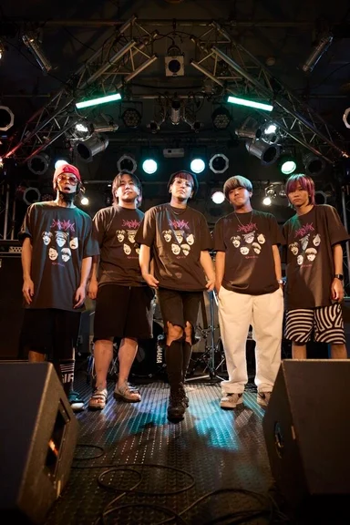 ライブハウスのステージに並ぶ5人。同じ顔のイラストをプリントしたTシャツを着ている