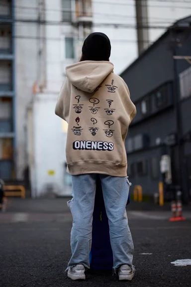 路上に立つ後ろ姿。顔のイラストとONENESSの文字が背面に入ったベージュのパーカー