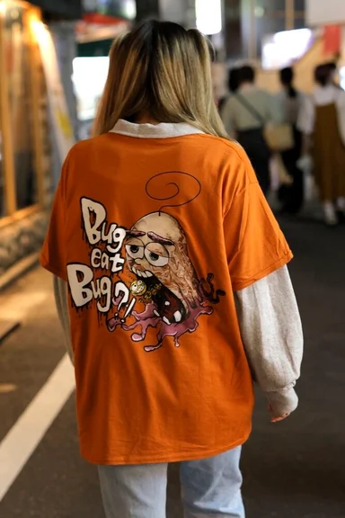 駅の通路を歩く後ろ姿。オレンジのTシャツの背面にキャラクターのイラスト