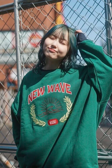 金網の前に立つモデル。NEW WAVEの文字と波を刺繍した緑のトレーナー