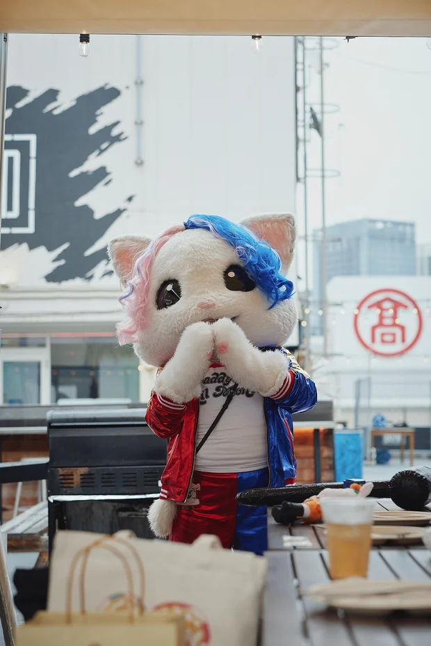 白猫レースクイーンのアップちゃん。赤と青のジャケットを着て、屋外のテラスで両手を口元に当てている