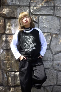 キャラクターと「That's Sick!」の文字を白でプリントした黒と白のラグランTシャツを着て、石垣の前に立つ女性