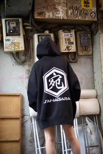 背中に「宛」の文字の六角形のロゴと「SANAGARA」を白でプリントした黒いパーカーを着た人の後ろ姿